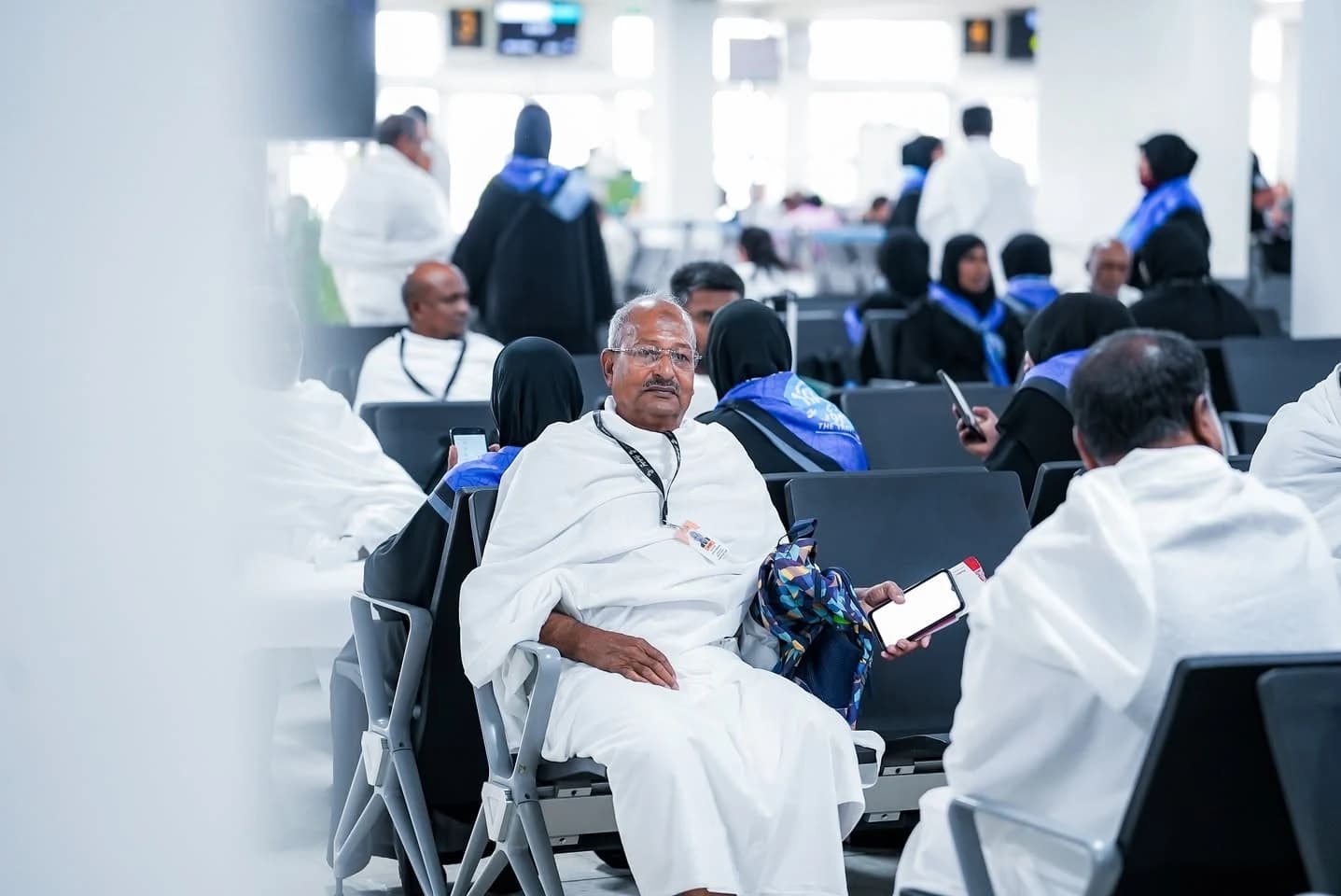 Pilgrims dressed in white ihram garments and black abayas sit in an airport departure lounge.