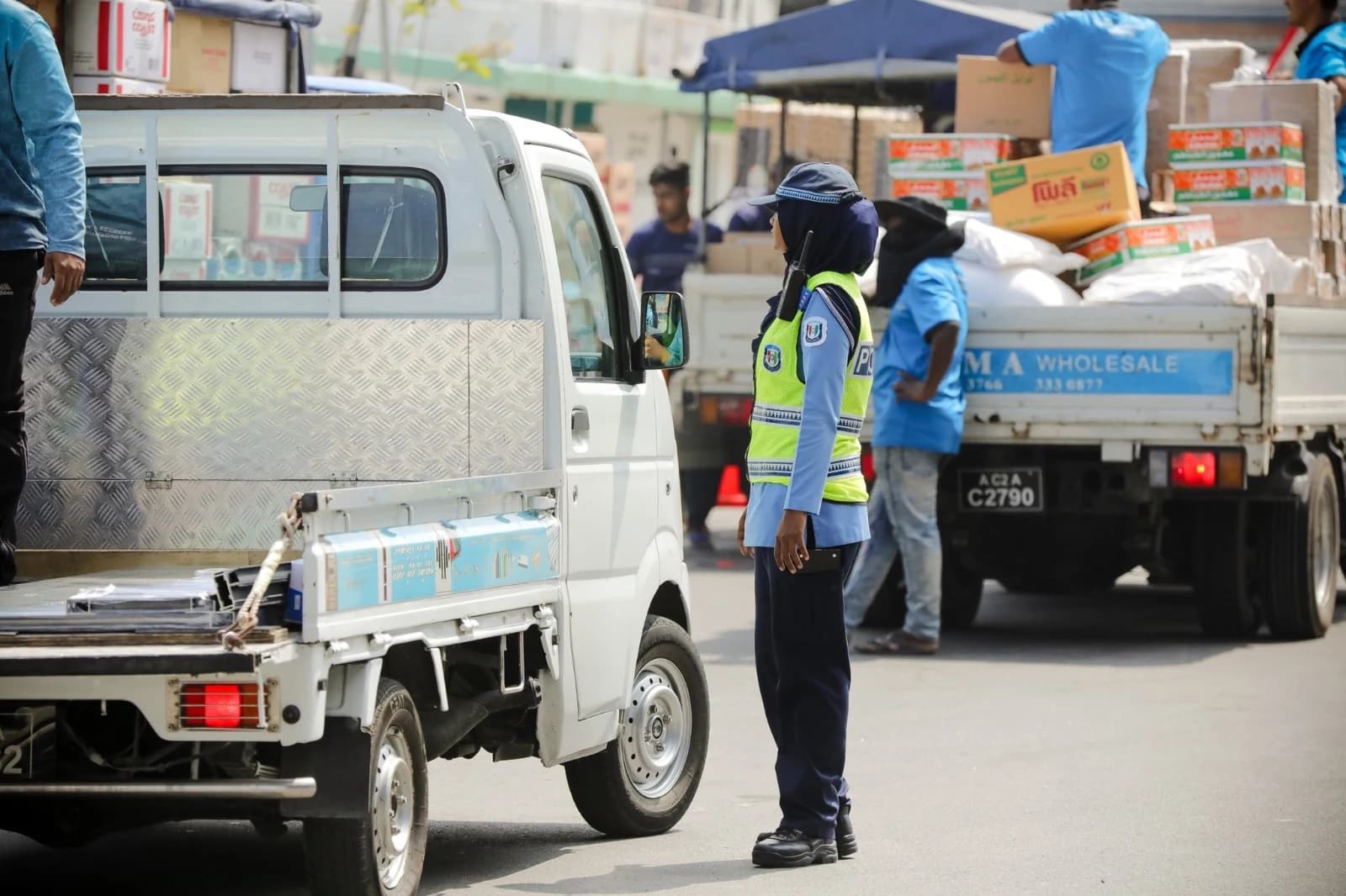 A police officer in a high-visibility vest stands on a Malé street between two small pickup lorries, one loaded with boxes and sacks by workers.