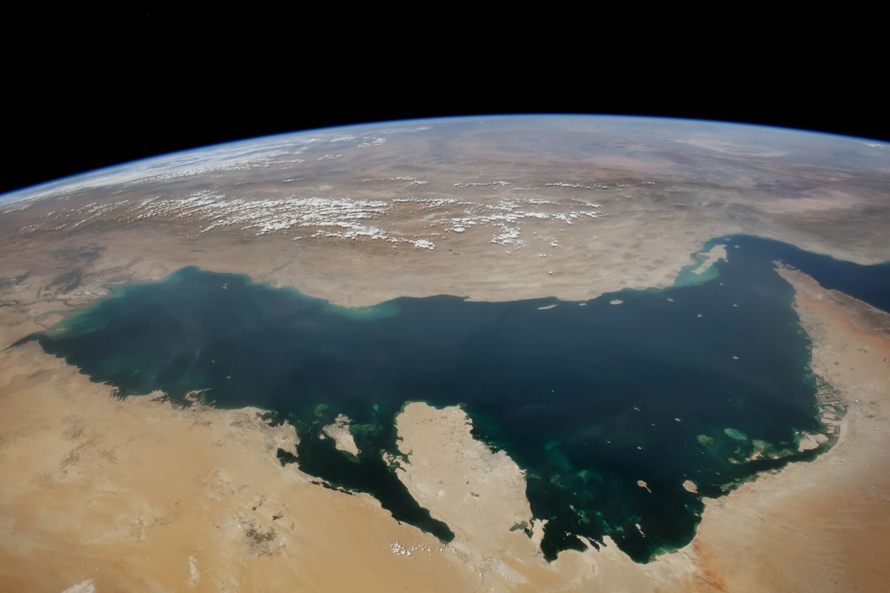 View from space of the Persian Gulf, a dark blue sea between pale desert coastlines, narrowing at right into the Strait of Hormuz under a curved horizon