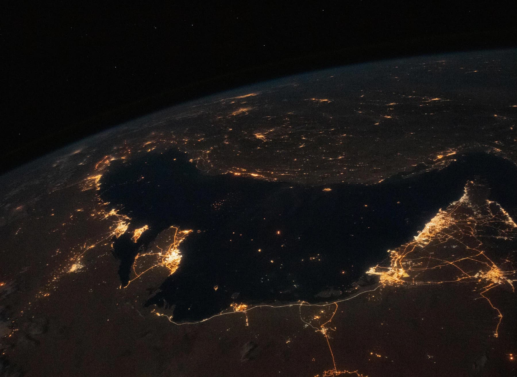 Night view from space of the Persian Gulf, a dark sea ringed by orange city lights, with scattered ship lights on the water and the Strait of Hormuz at right