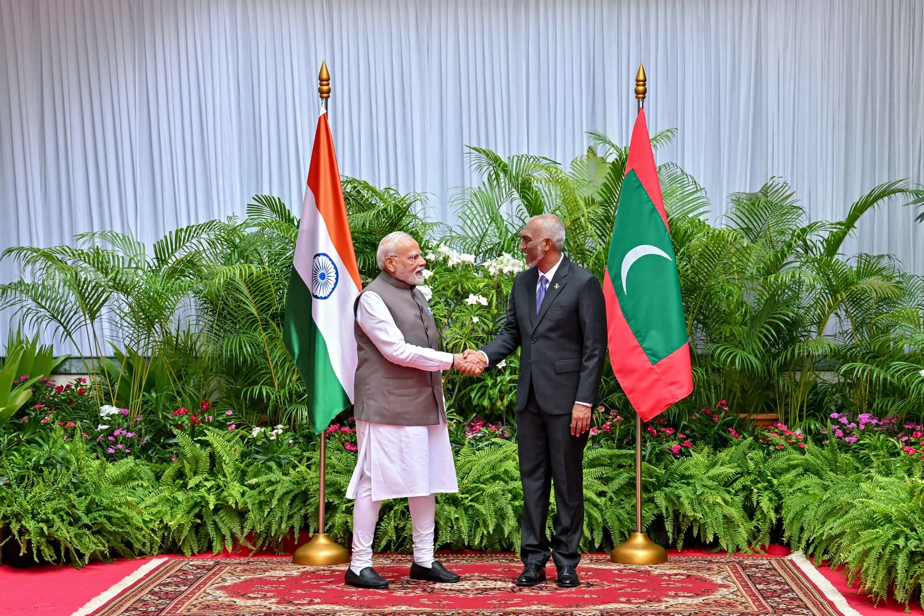 Narendra Modi, in a grey waistcoat, shakes hands with Mohamed Muizzu, in a dark suit, between the flags of India and the Maldives in front of a bank of tropical plants.