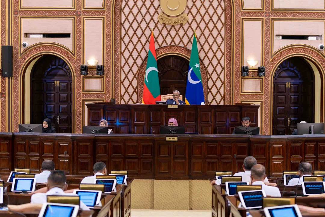 The People's Majlis chamber, with the presiding officer seated between the national flag and the Majlis flag and members at their desks in the foreground.