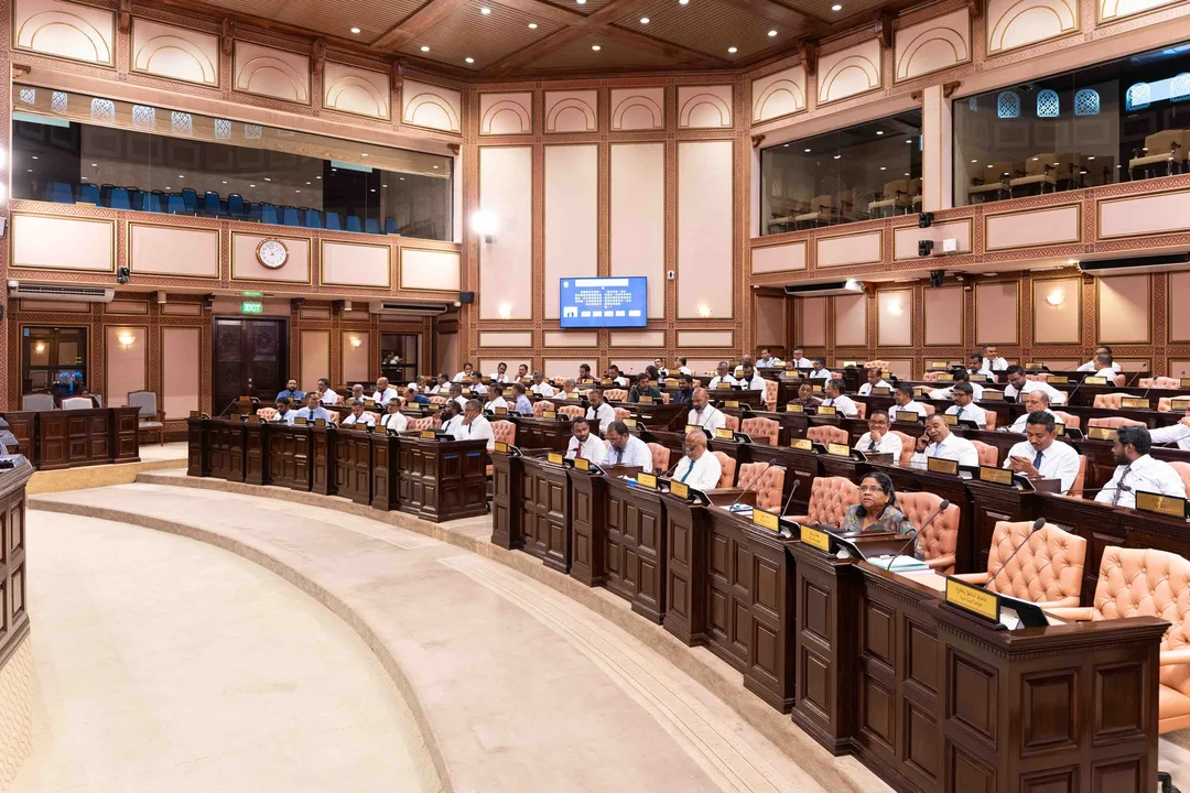 Members of the People's Majlis at their desks in the chamber, seen from the side, below the public galleries and a wall screen showing the chamber's seating plan.