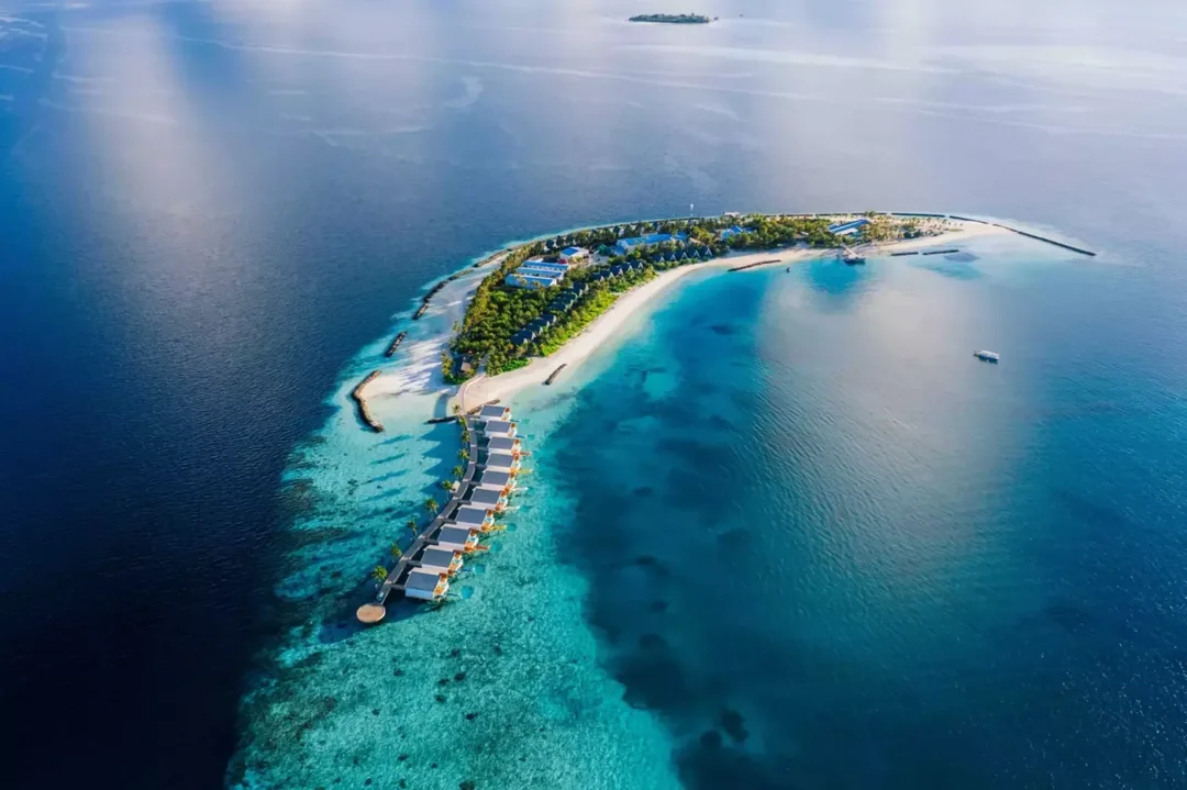 Aerial view of a curved resort island with white beaches, trees and villas, rock breakwaters lying off its shore, and a row of overwater villas on a jetty running out over the reef.
