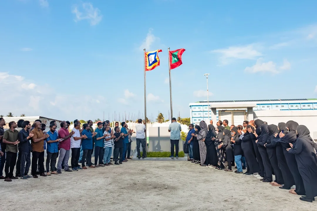 Staff stand in two lines applauding as the national flag and the STELCO flag fly on poles in front of a utility building under a blue sky.